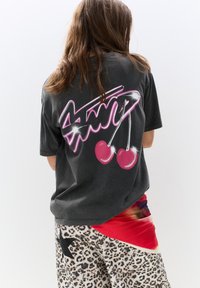 T-shirt oversized gris avec un graphique en texte néon rose et des cerises au dos ; porté avec un short à motifs et une pièce rouge superposée.