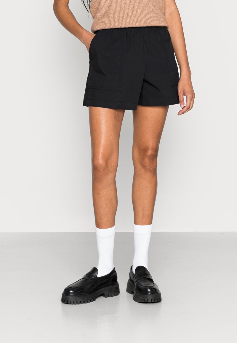 Femme portant un short noir taille haute avec poches avant, des chaussettes blanches, des mocassins noirs épais, et un haut beige sans manches rentré.