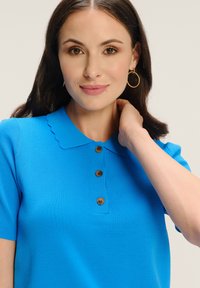 Jasnoniebieska koszulka polo z krótkim rękawem, z falbanym kołnierzykiem i trzema brązowymi guzikami. Model pozuje z ręką na szyi, prezentując fakturowany materiał.
