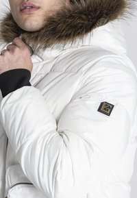 Giacca puffer bianca con cappuccio foderato in pelliccia marrone, polsini neri a costine e una toppa con logo sulla manica. Presenta tasche con zip.