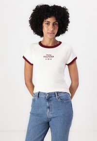 Bela majica s bordo rebrastim ovratnikom i rubovima rukava, s "Tommy HILFIGER" logotipom u crvenom tekstu. Uparena s plavim trapericama.