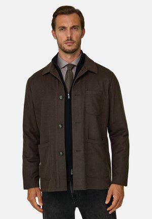 Homme portant une veste marron foncé boutonnée par-dessus une couche noire zippée, chemise grise et cravate marron assortie, debout devant un fond uni.