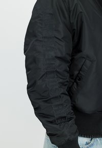 Czarny rękaw kurtki z pionowo wyhaftowanym "Alpha Industries" czarną, dopasowaną nitką, noszony z jasnoniebieskimi dżinsami.