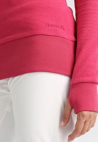 Sudadera rosa ajustada con puños acanalados y dobladillo ancho. Presenta una sección superior texturizada y un logo "Bench." en relieve. Pantalones blancos debajo.