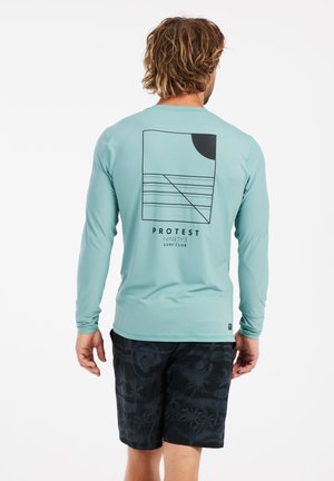 Camicia azzurra a maniche lunghe con stampe geometriche sulla schiena, abbinata a pantaloni corti neri con un motivo di palme, mostrando una vestibilità rilassata.