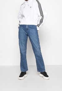 Person som bär en vit Adidas-hoodie med svarta ränder, blå raka jeans och svarta-vita chunky sneakers, stående mot en vit bakgrund.