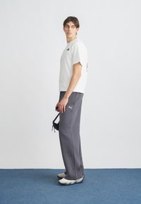 Λευκό βαμβακερό T-shirt με λογότυπο στο στήθος, συνδυασμένο με γκρι φόρμα με λογότυπο στον μηρό. Το μοντέλο φοράει αθλητικά παπούτσια.