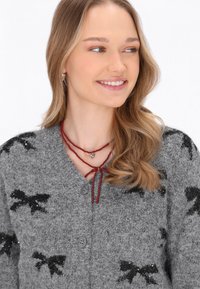 Šedý pletený cardigan s černými vzory mašlí, s knoflíkovým zapínáním a měkkou texturou. Modelka nosí vrstvený červený náhrdelník.