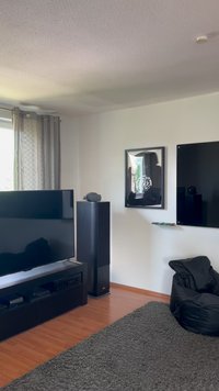 Televisor negro sobre un soporte elegante, al lado de un altavoz alto. Alfombra gris oscuro, suelo de madera clara y un puff negro. Arte en la pared enmarcado.