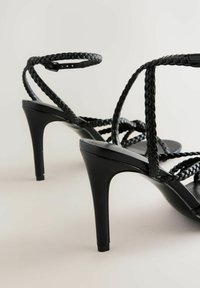 Schwarze High-Heelsandalen mit geflochtenen Riemen, die einen schlanken Absatz und eine glatte, glänzende Sohle aufweisen. Minimalistisches Design mit verstellbarem Knöchelriemen.