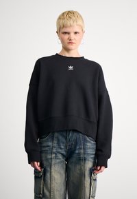 Juoda per didelė medvilninė džemperis su apvaliu kaklu ir mažuoju baltu Adidas logotipu ant krūtinės. Dėvima su mėlynais kargo džinsais.