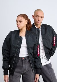 Jachete negre tip bomber cu textură perforată, manșete cu arcuri și fermoare. O jachetă are un etichetă roșie cu text alb. Imaginea prezintă un set asortat cu pantaloni gri.
