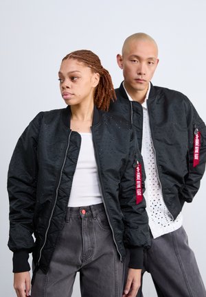 Jachete negre tip bomber cu textură perforată, manșete cu arcuri și fermoare. O jachetă are un etichetă roșie cu text alb. Imaginea prezintă un set asortat cu pantaloni gri.
