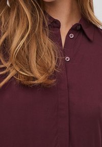 Camicia bordeaux con colletto, caratterizzata da due bottoni e una superficie tessile liscia, parzialmente visibile contro i capelli chiari.