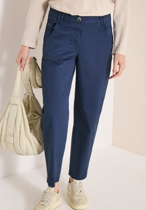 Kobieta w granatowych dopasowanych spodniach, beżowych trampkach i beżowej bluzce z długim rękawem, trzymająca beżową torebkę na ramieniu.