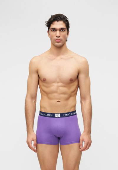 Homme musclé aux cheveux foncés portant un boxer violet Polo Ralph Lauren, debout devant un fond gris clair uni.