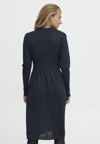 Abito blu navy realizzato in tessuto testurizzato con maniche lunghe, vita svasata e finitura leggermente lucida, che arriva sotto il ginocchio.