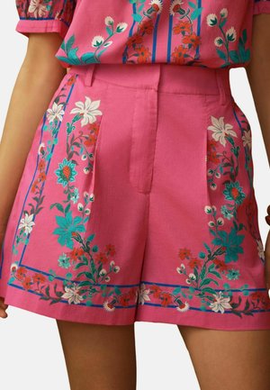 Pantaloni scurți roz cu talie înaltă, cu modele florale albe, teal și roșii de-a lungul părților laterale și tivului, purtați cu un top floral asortat.