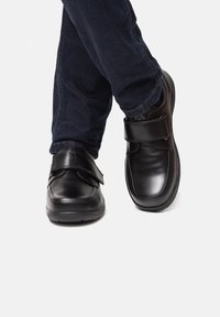 Scarpe in pelle nera con punta arrotondata e cinturino in velcro regolabile, abbinate a jeans blu scuro, esposte in posizione verticale.