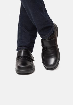 Black leather shoes with a rounded toe and adjustable Velcro strap, paired with dark blue jeans, displayed in a standing position.