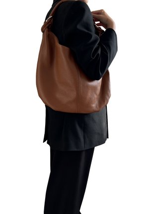 Persona vestita con un completo nero che porta una grande borsa a tracolla in pelle marrone texturizzata sul lato destro.