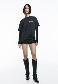 Črna prevelika majica z belim grafičnim motivom, nošena čez oprijet zgornji del iz čipke, kombinirana s črnimi kratkimi hlačami s sequini in črnimi škornji do kolen.