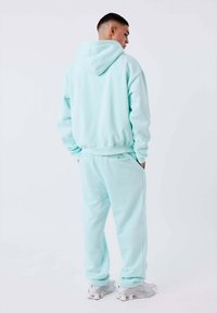 Giovane uomo in piedi di spalle, che indossa un felpa azzurra chiara e pantaloni della tuta abbinati, con sneakers bianche su sfondo bianco.