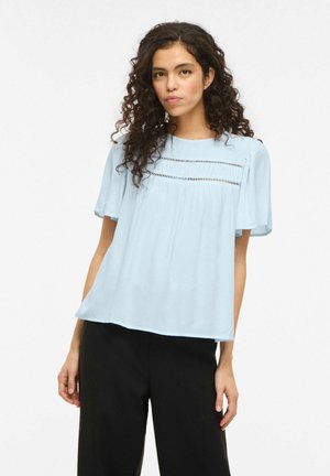 Kvinde med krøllet hår iført en lyseblå kortærmet bluse med horisontale udskæringsdetaljer og sorte bukser, stående mod en hvid baggrund.