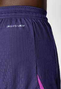 Zoom na temno purpurne teksturirane atletske hlače slogom "DRI-FIT ADV" in rožnato notranjo oblogo, ki je vidna ob stranski zarezi.