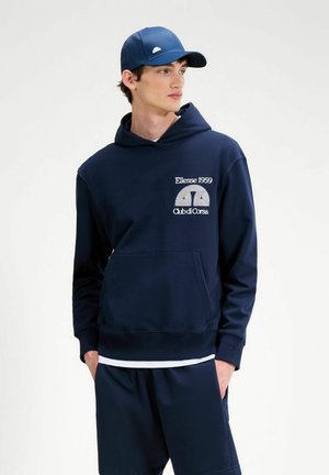 Giovane uomo che indossa una felpa con cappuccio blu navy e pantaloncini con il logo "Ellesse 1959 Club di Corsa", e un cappellino da baseball blu navy coordinato, guarda di lato.