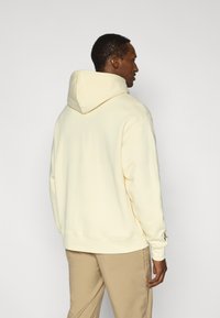 Πουλόβερ με κουκούλα σε ανοιχτό κίτρινο, κατασκευασμένο από απαλό ύφασμα. Διαθέτει κουκούλα με κορδόνια, ριμπ μανσέτες και λεπτομέρεια με λογότυπο στο μανίκι.