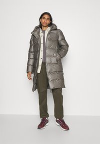 Lang grå pufferjakke med hette, med en quiltet design og to sidelommer. Kombinert med olivengrønne bukser og maroon sneakers.