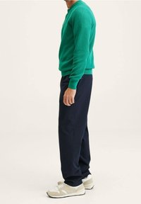 Mann, der seitlich steht und einen grünen Pullover, dunkelblaue Hosen und beige-weiße Sneakers trägt, vor einem schlichten hellen Hintergrund.