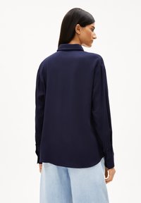 Marineblaue Langarmhemd mit Knöpfen und Kragen. Glattes Material, dezenter Glanz und abgerundeter Saum. Lockere Passform, getragen über hellblauen Hosen.