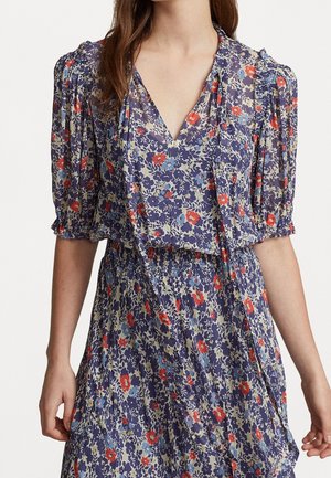 Femme portant une robe fleurie avec des manches bouffantes, un décolleté en V et une taille smockée en bleu, rouge et crème.