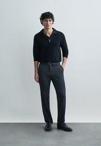 Black zippered sweater, tailored gray pants, black belt and shoes. Model stands against a neutral background, presenting a modern, fitted look.