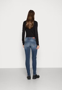 Slim-fit blå jeans i denim med medelblå tvätt, med fem fickor och en brun läderpatch på bakre midjan. Svarta ankelstövlar bärs.