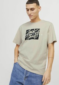 Beige T-Shirt mit kurzen Ärmeln und einem schwarz-weißen Grafiklogo, das zentral auf der Brust platziert ist. Weicher Stoff, lässige Passform und Rundhalsausschnitt.