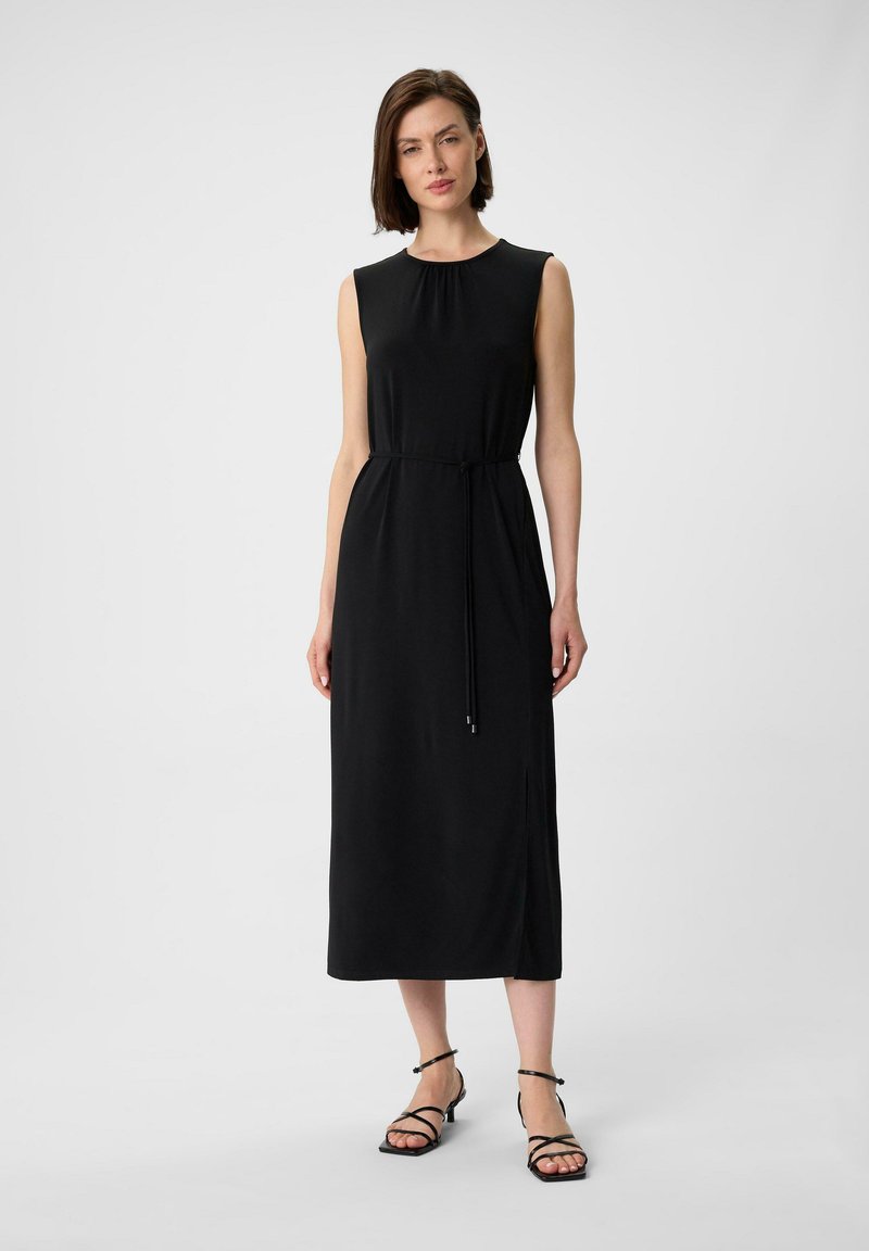 Schwarzes ärmelloses Kleid mit einem runden Ausschnitt, Taillendetail mit Bindegürtel und seitlichem Schlitz; glattes Material mit fließender Silhouette. Schwarze Sandalen mit Riemchen.