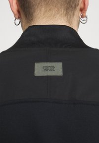 Svart sweatshirt med ribbad krage, som har en rektangulär grön etikett med text på baksidan, slätt tyg och kontrasterande texturer.
