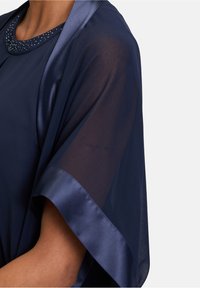 Navy blue top with sheer fabric and satin trim. Features a beaded neckline and a subtle handwriting detail on the sleeve.