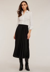 Camicia bianca con bottoni e maniche lunghe abbinata a una gonna nera plissettata di media lunghezza con vita elasticizzata e stivali neri fino al ginocchio.