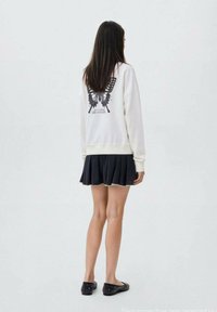 Krémová mikina s grafickým motivem motýla na zadní straně, doplněná černou plisovanou sukní. Černé baleríny se cvočky dokončují outfit.