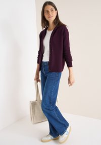 Cárdigan de punto burdeos con cierre de botones sobre una camiseta blanca, combinado con jeans azules acampanados y zapatillas blancas, llevando un tote crema.