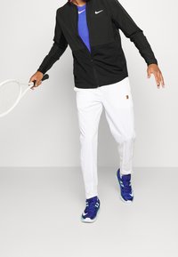 Black zip-up jacket over a blue shirt, white pants, and blue-green tennis shoes. Holds a tennis racket. Simplistic design and modern fit.