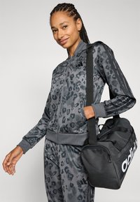 Grå leopardmönstrad träningsdräkt med en zip-up jacka och matchande byxor. Svart sportväska med vitt logo och stadiga remmar.