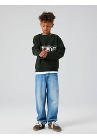 Mørkegrønn genser med et retro TV-grafikk, kombinert med lyseblå vide jeans og brune joggesko med hvite skolisser.