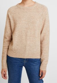 Pullover color beige chiaro realizzato in un morbido tessuto testurizzato. Presenta un collo rotondo, maniche lunghe e polsini e orlo a coste. Indossato sopra un paio di jeans.