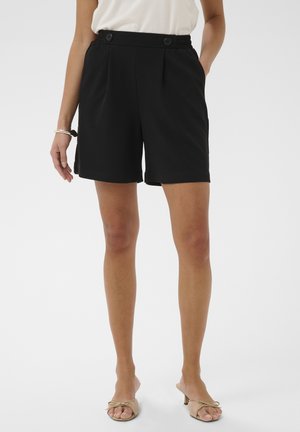 Mujer con shorts negros de cintura alta con detalles de botones, blusa blanca, tacones beige abiertos en los dedos y pulsera plateada, de pie sobre fondo blanco.