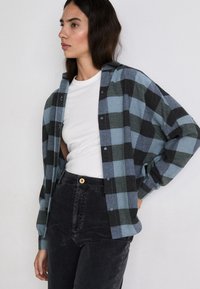 Camicia di flanella a pattern scozzese blu e nero, tessuto leggero, con bottone frontale, vestibilità rilassata, indossata sopra una maglietta bianca e jeans neri.